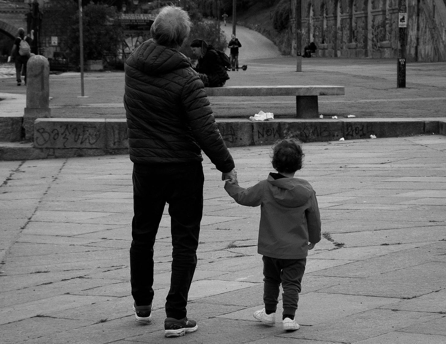 Older man holds the hand of a child. They look across a road together, backs to the camera.