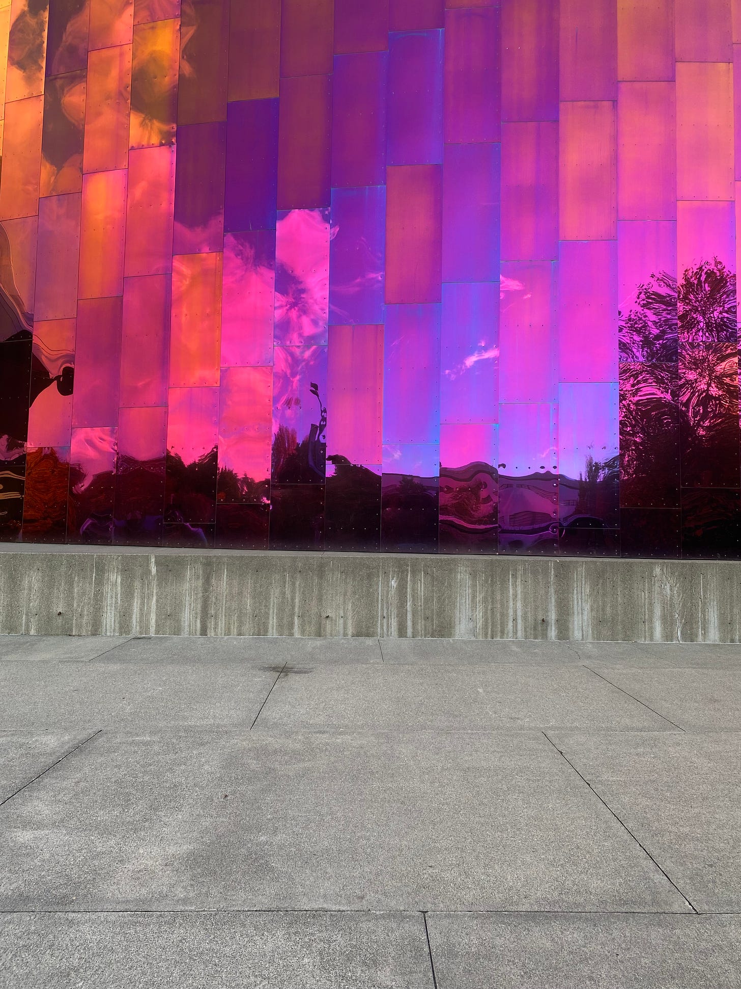 A building with reflective pinkish-purple panels, showing the sky and some trees. The sidewalk and a concrete ledge are in the bottom half of the photo.