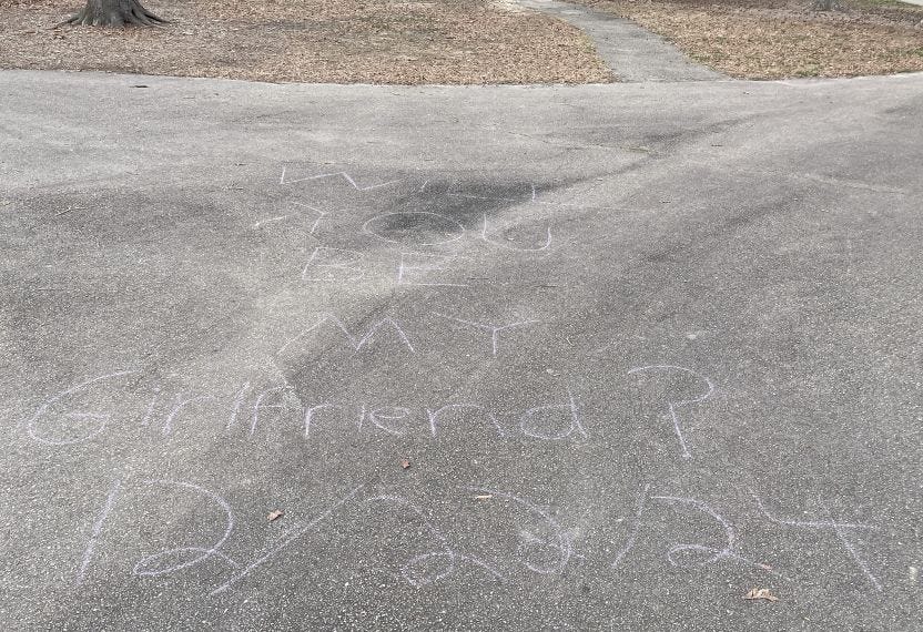 blacktop with words in pale chalk letters: WILL YOU BE MY Girlfriend 12/22/24; a border of mulch in a treed area visible along top edge of photo