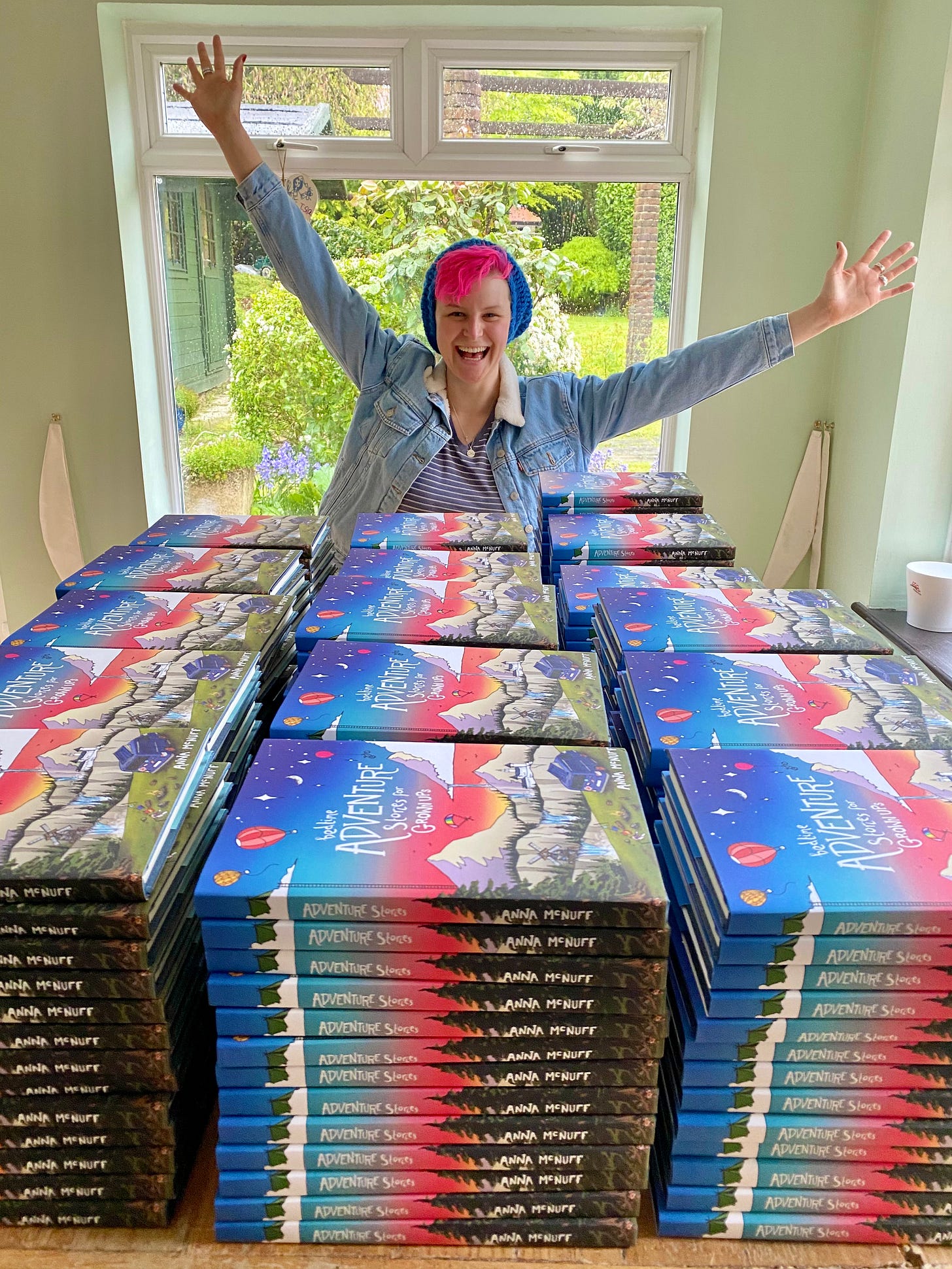  Author and adventurer Anna McNuff stands beaming behind a table stacked high with colourful copies of her book "Adventures Stories for Rebel Girls." Arms raised in triumph, she sports a denim jacket, pink hair, and a blue knit hat. Behind her, a lush garden peeks through the rain-flecked windows.