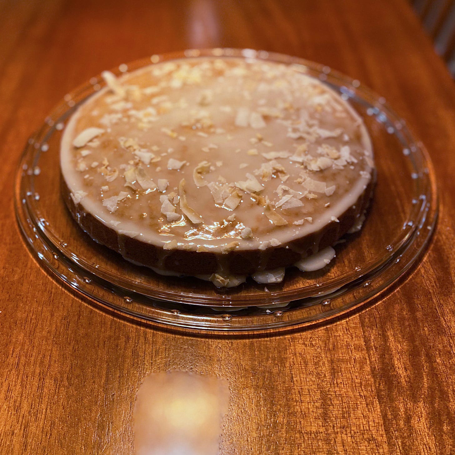 A golden cake in a white glaze that has dripped over the edges, onto a glass cake plate. Large flakes of coconut are sprinkled over the top.
