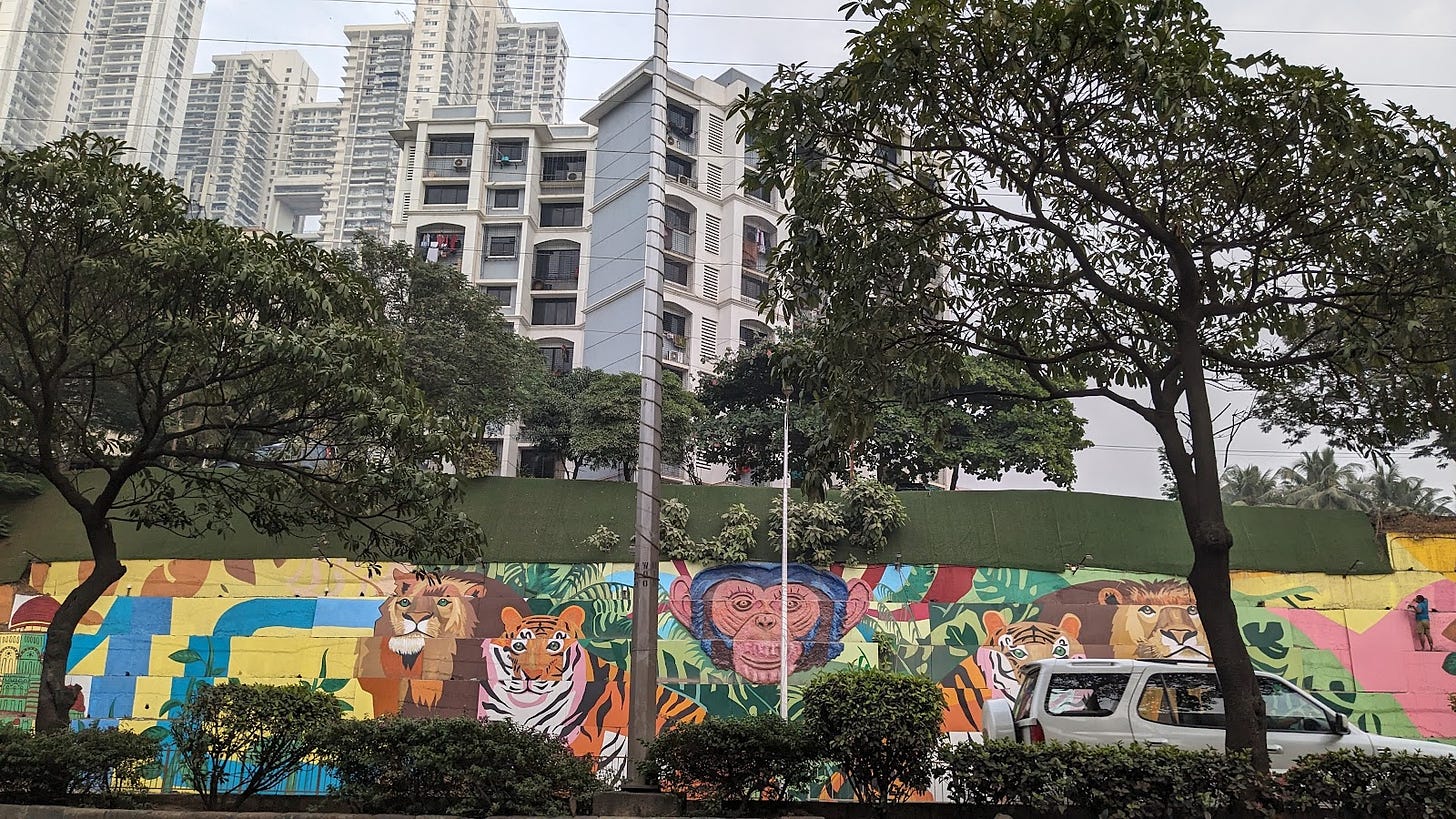 Urban street showing a colorful mural of a jungle scene painted with lions, tigers, and monkeys. High-rise buildings rise up behind the mural.