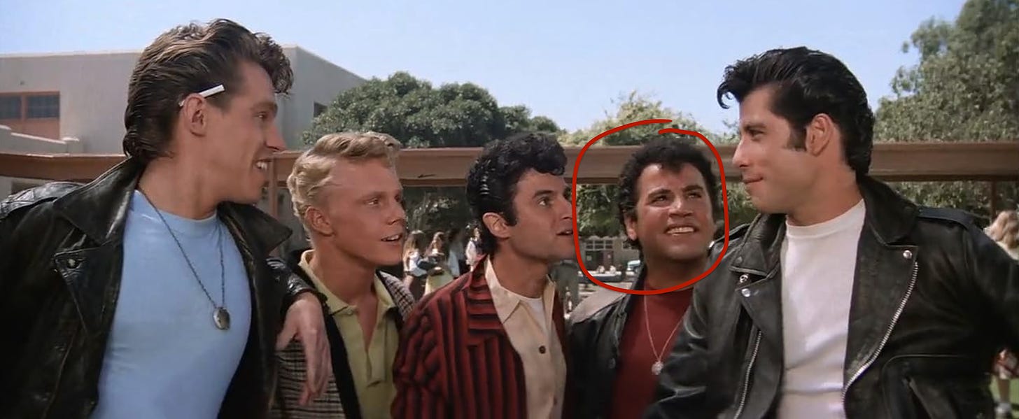 A screen capture of the movie Grease. The T-Birds, five teenager greasers, stand beside each other outdoors on a sunny day. A red circle is around Sonny's face.