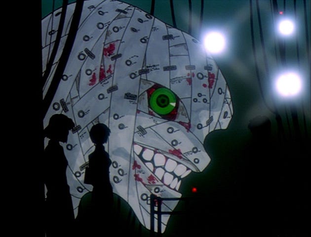 A side view of EVA-01 without its purple armor plating. It is a massive human-like head with bandages stapled onto it and striking green eyes, staring directly at the viewer. In front, the silhouette of Ritsuko and Maya, two NERV scientists, i visible, as well as the silhouette of some wiring and machinery. The entire scene is dimly lit with artificial light.