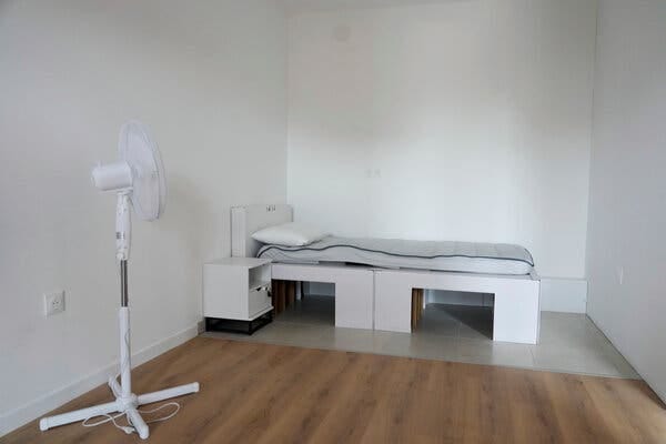 A small bedroom has white walls and a wooden floor. A white bed is in the corner and there is a white fan on a stand near the wall. 