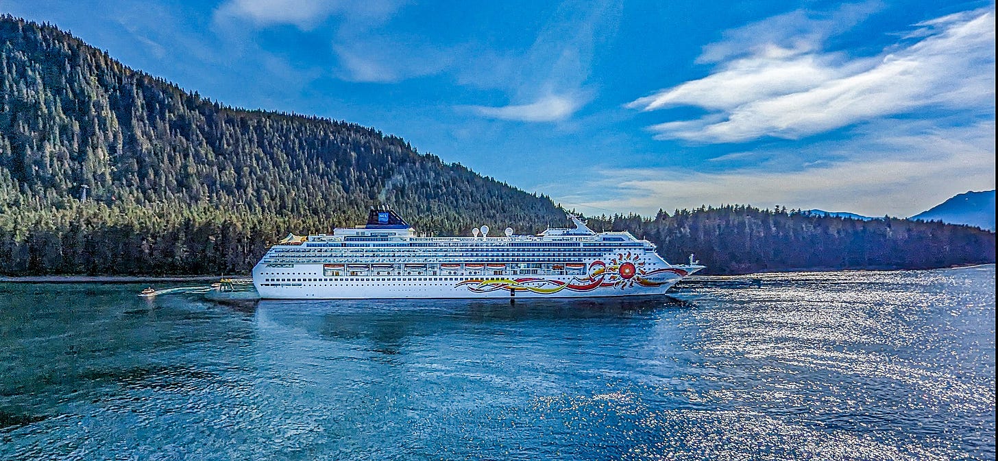A cruise ship moored in front of tree covered mountains under a blue sky. 