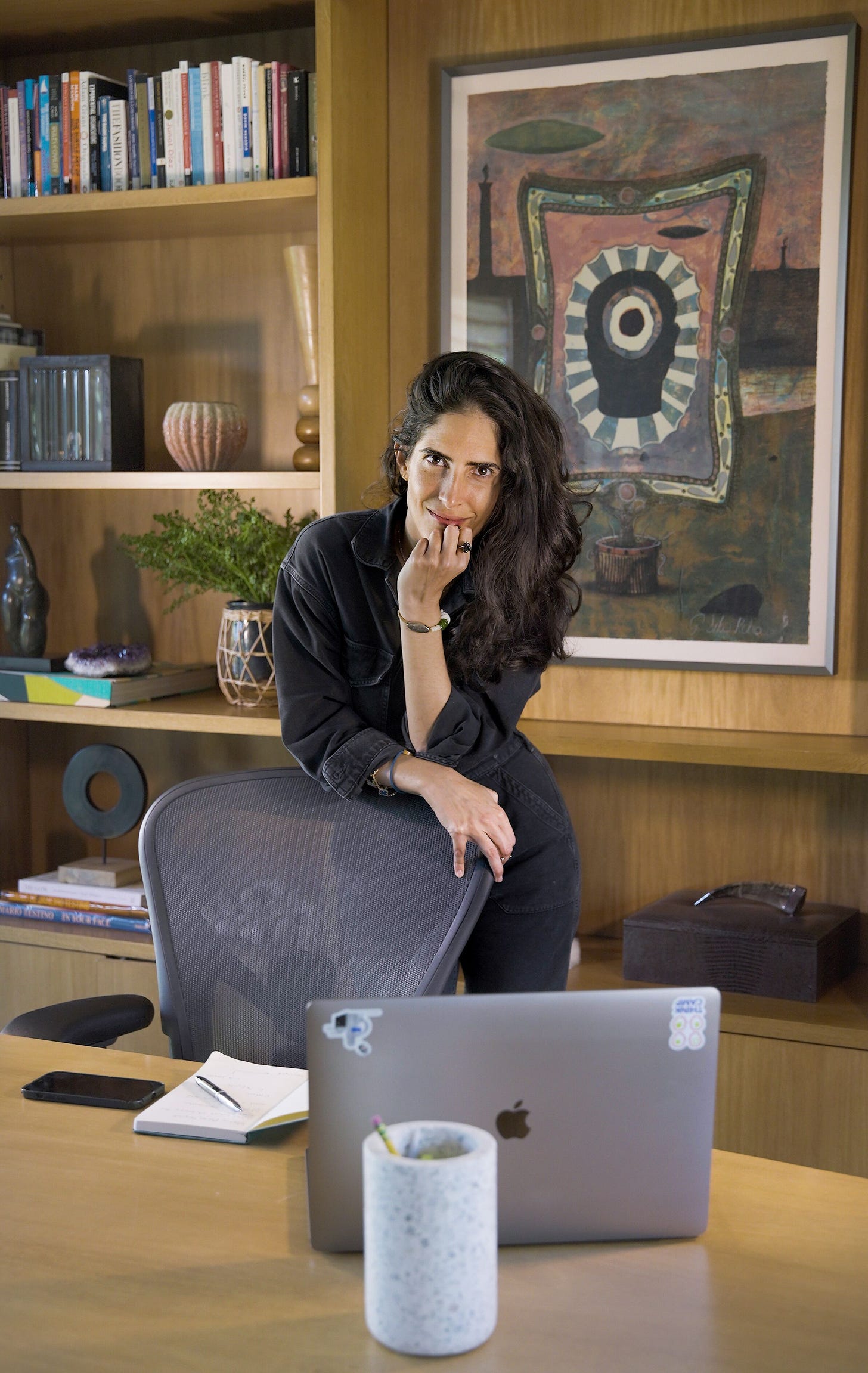 Sari is standing in an office, leaning over a grey Herman Miller office chair with her right hand resting on the left corner. She’s wearing a black shirt and black jeans, and her long black, wavy hair is draped round her left shoulder. Behind Sari is a large portrait abstract painting, to the right of which is a wooden bookshelf featuring a range of books, ceramics, and sculptures, all carefully placed.