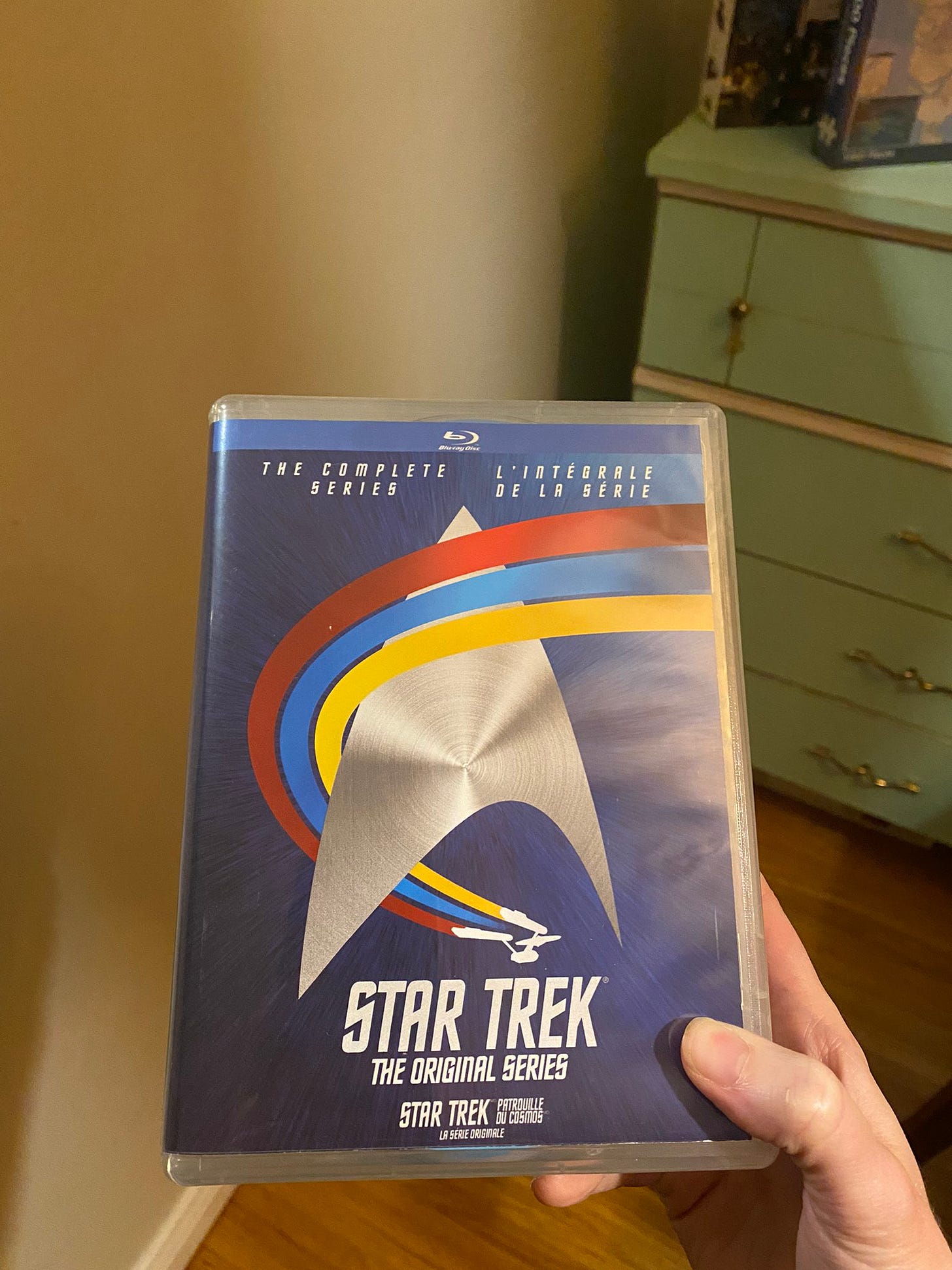 My hand holding a blu-ray case for the complete series of Star Trek: The Original Series. It’s a dark blue case with a large silver Starfleet delta, the Enterprise in the background trailing red, blue, and yellow from around the front of the delta logo. The set is bilingual with English and French because I live in Canada where everything is like that.