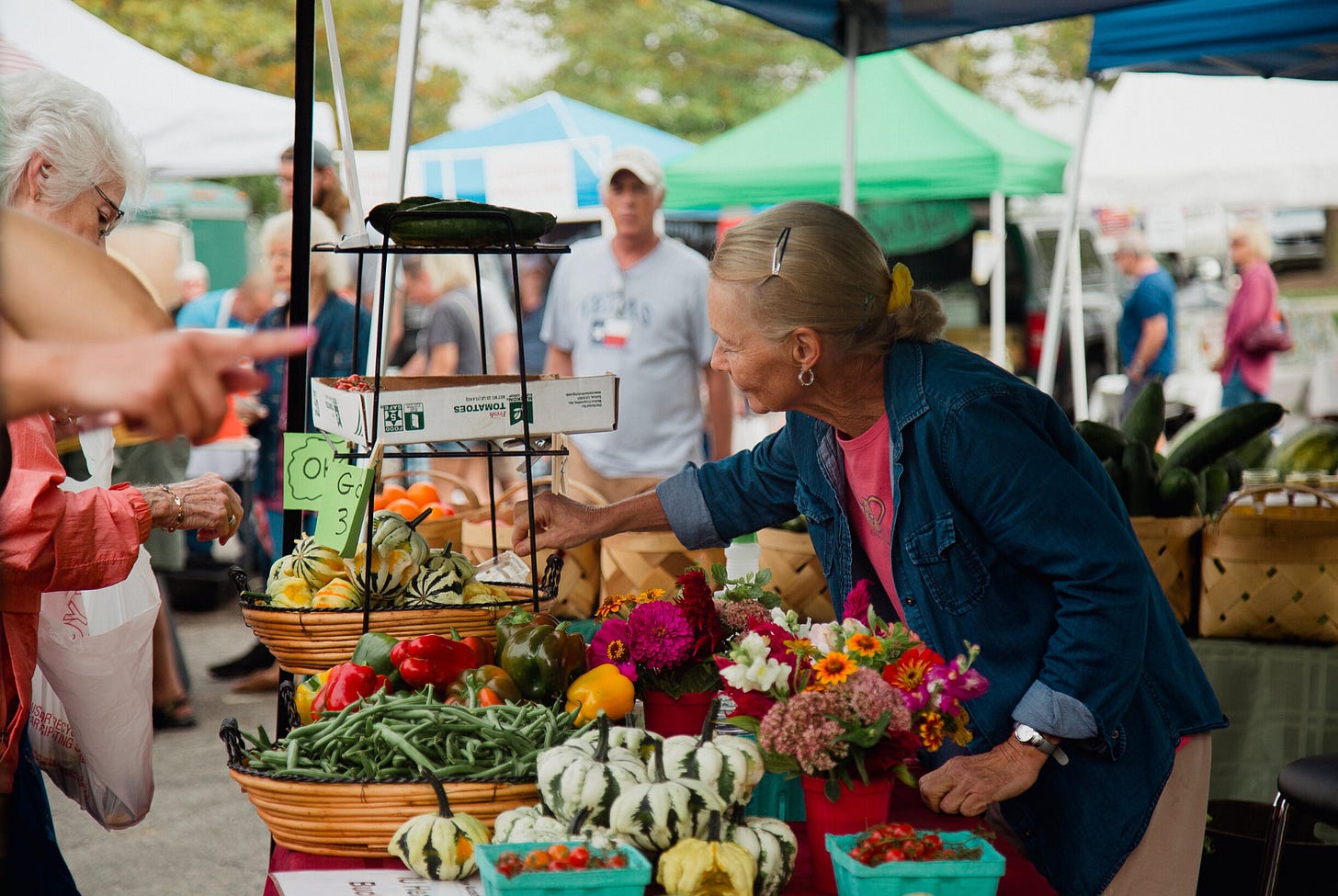 A woman leans over a table at a open-air market, the table filled with all kinds of fresh vegetables, fruit, and flowers. Other people can be seen, blurry, in the background.