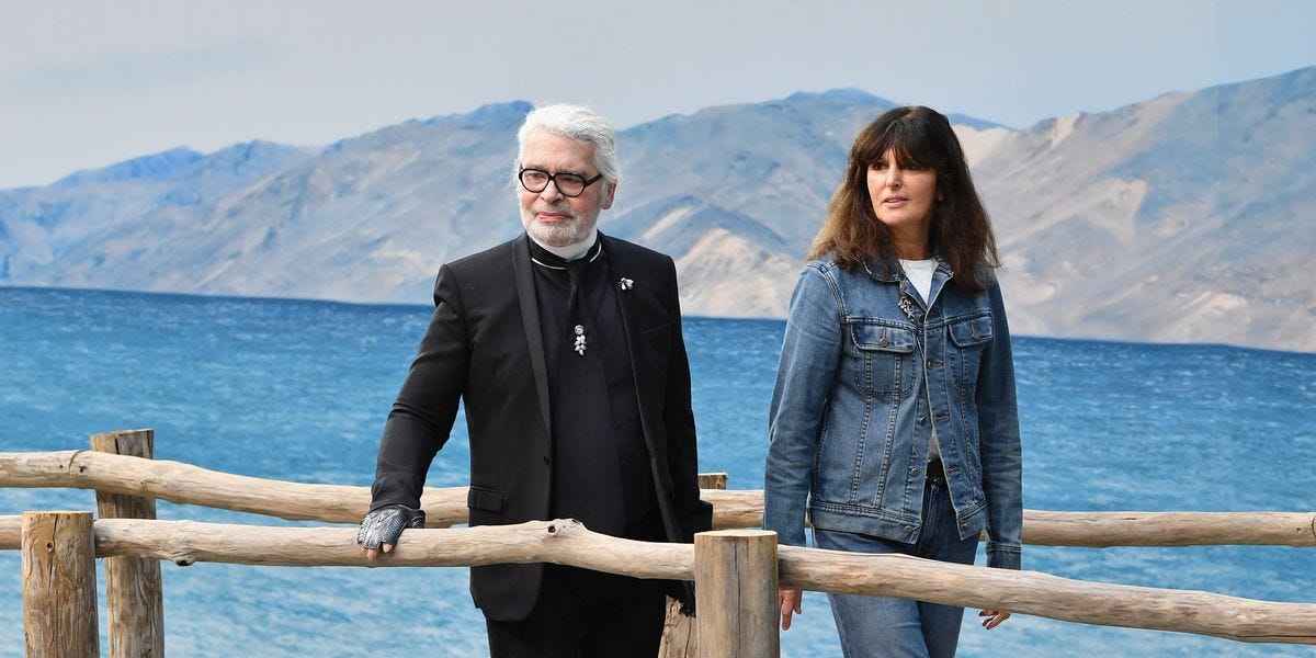 An elderly main dressed in a black suit with black glasses stands next to a mature woman with shoulder-length brown hair and a denim jacket over a white t-shirt worn with jeans. 