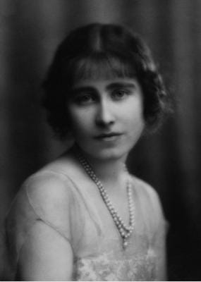 Black and white, head-and-shoulders photograph of Elizabeth Bowes Lyon, seen from the side with her face turned to face the viewer. She is wearing a light, lacy dress with short sleeves, and two strands of pearls. Her dark hair is parted in the middle, with a fringe.