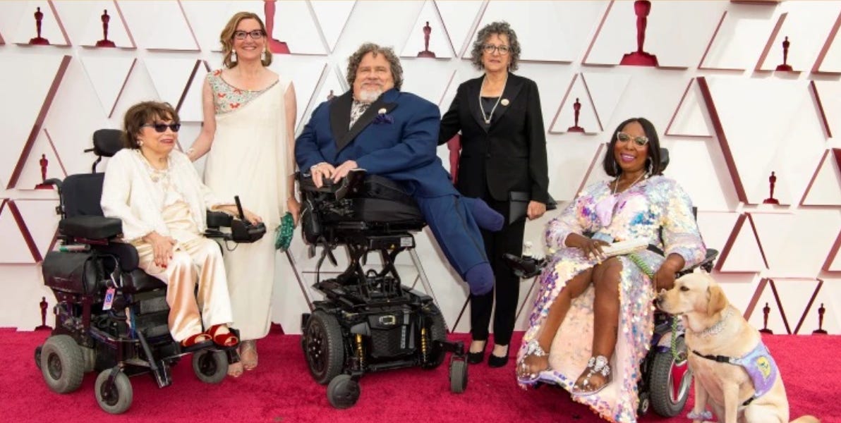 Foto no tapete vermelho de parte do elenco do documentário Crip Camp: três pessoas em cadeiras de rodas motorizadas posam para fotografia; um cão de serviço está no canto direito da imagem. 