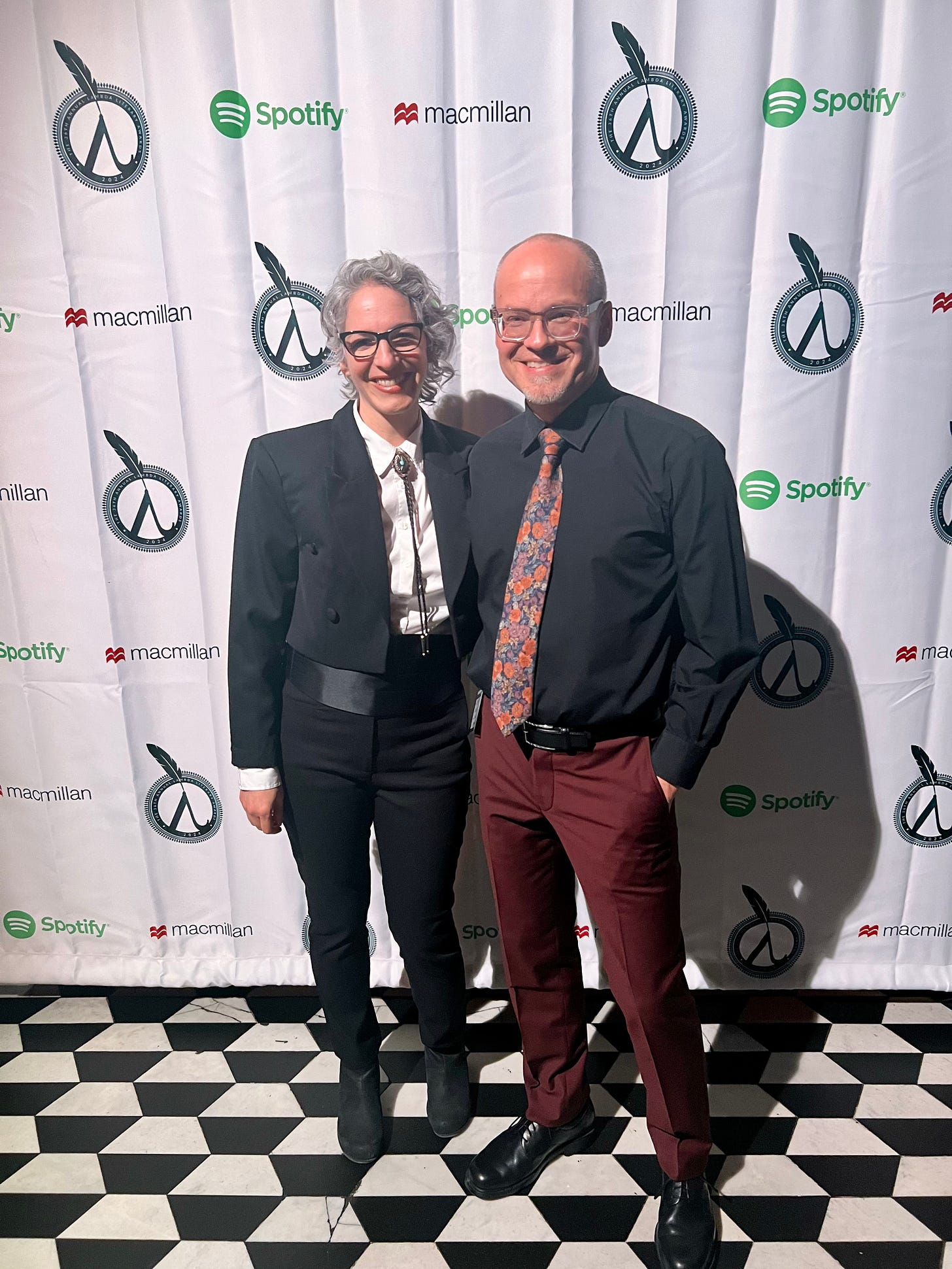 Me posing with my husband Chris in front of the step and repeat at the Lammy Awards. I have short grey wavy hair and am wearing a tux with a cropped jacket and a bolo tie. Chris is bald and wearing a black shirt with a floral tie and dark red pants.