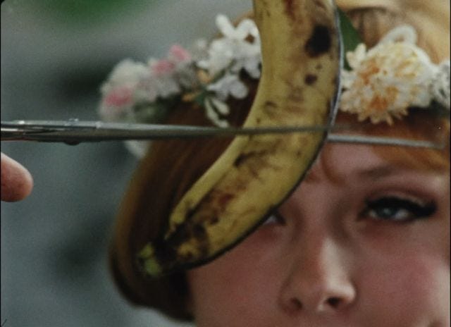 imagem colorida; uma adolescente loira, de cabelos curtos e usando uma coroa de flores, está cortando uma banana com uma tesoura. ela tem um olhar muito travesso e usa um delineador grosso nos olhos
