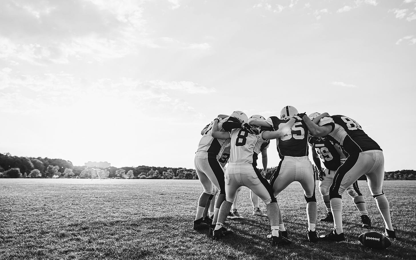 Team of football players huddle on an empty grass field. The sun sits on the horizon bathing the field in glorious light.