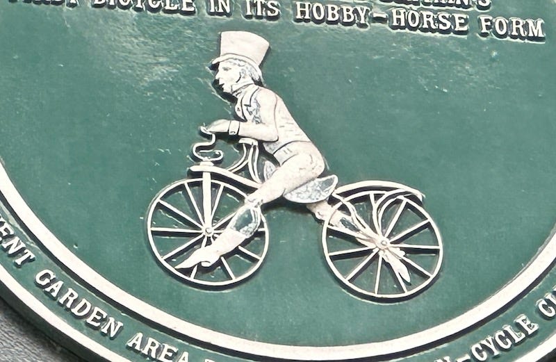 detail of a green plaque showing a hatted man on a hobby horse cycle