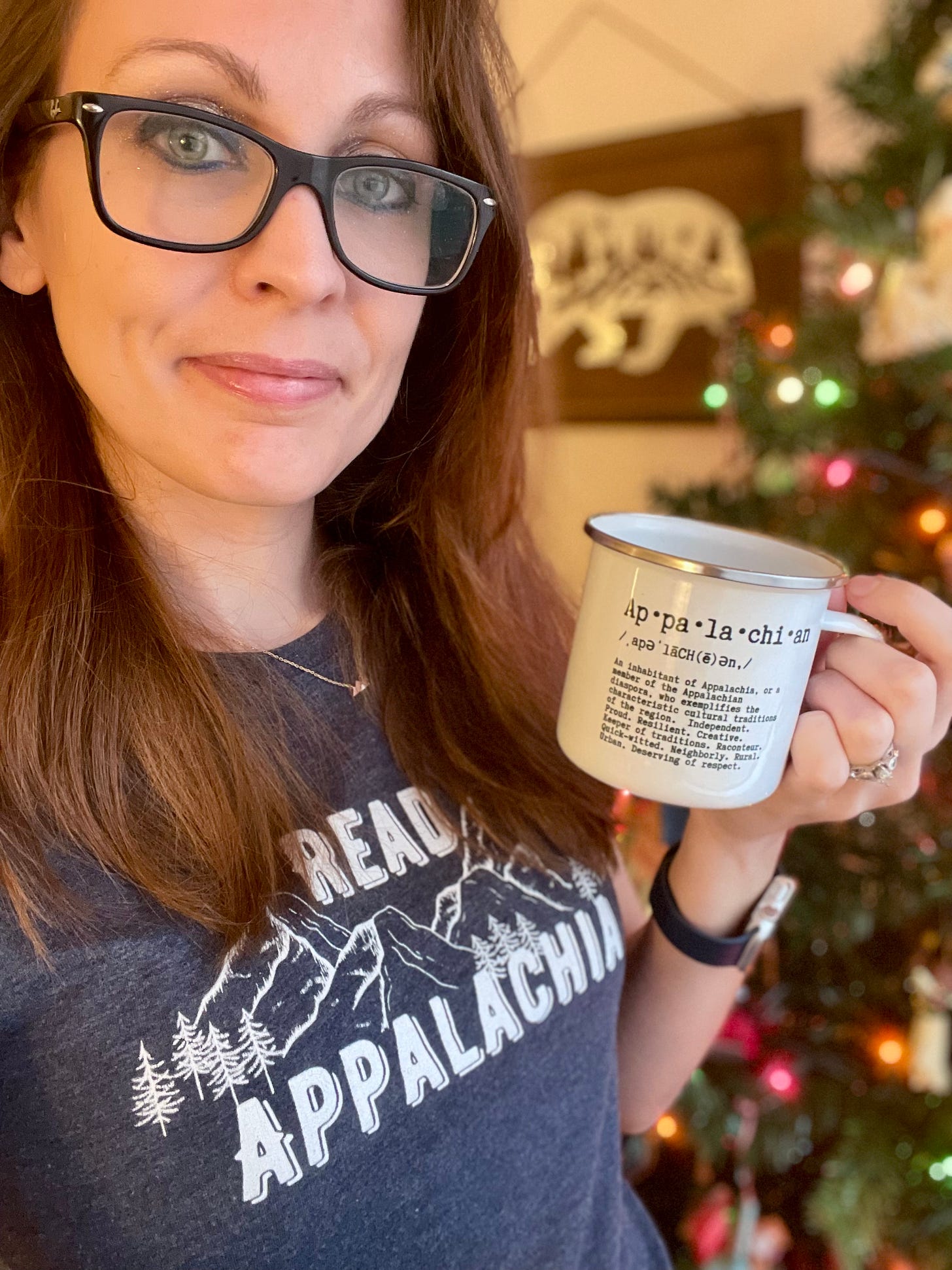 a photo of Kendra, a white woman with brunette hair wearing black glasses. She's wearing a Read Appalachia t-shirt and holding a mug that says Appalachia