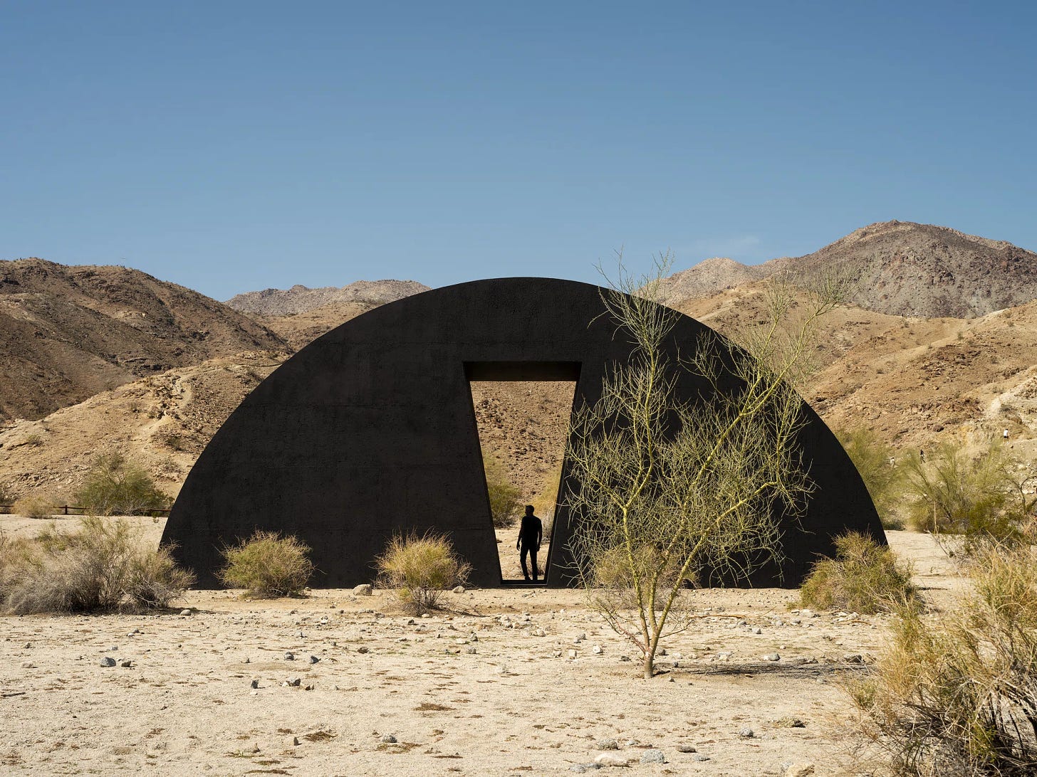 A black scuplture the shape of a half moon emerging from the desert landscape with a person walking through a portal, or path, or keyhole cut out the shape of a long upside down triangle with the tip cut off.