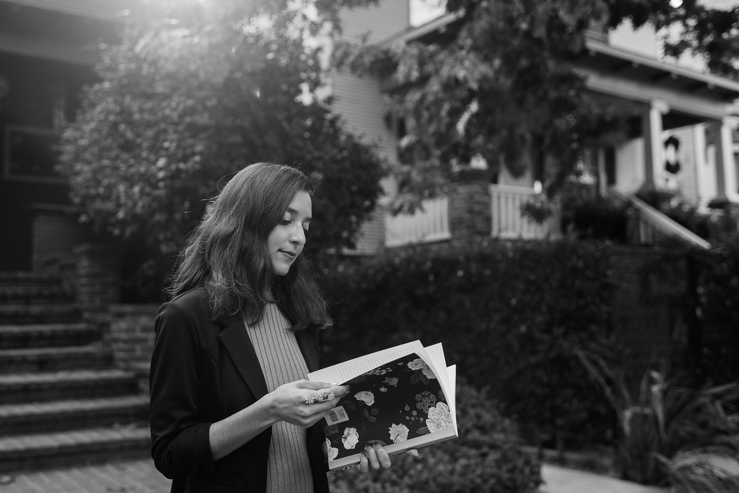 A black-and-white photo of a brown woman with short dark hair. She holds a floral black notebook and flips through its pages. The woman wears a black blazer and light dress. Behind her are trees and homes.