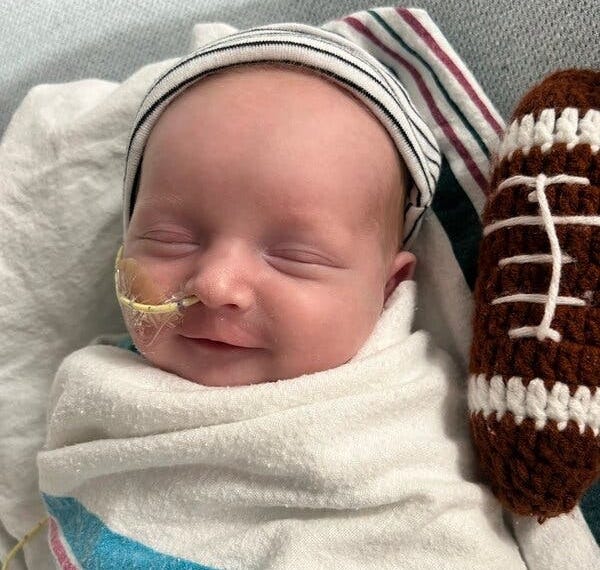A close-up portrait of newborn KJ, who has a tube coming out of his nose and a tiny felt football next to him. He is swaddled tight and appears to smile slightly with his eyes closed.