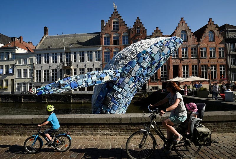 A whale statue made of pieces of plastic in a European square, with kids and a mom biking by