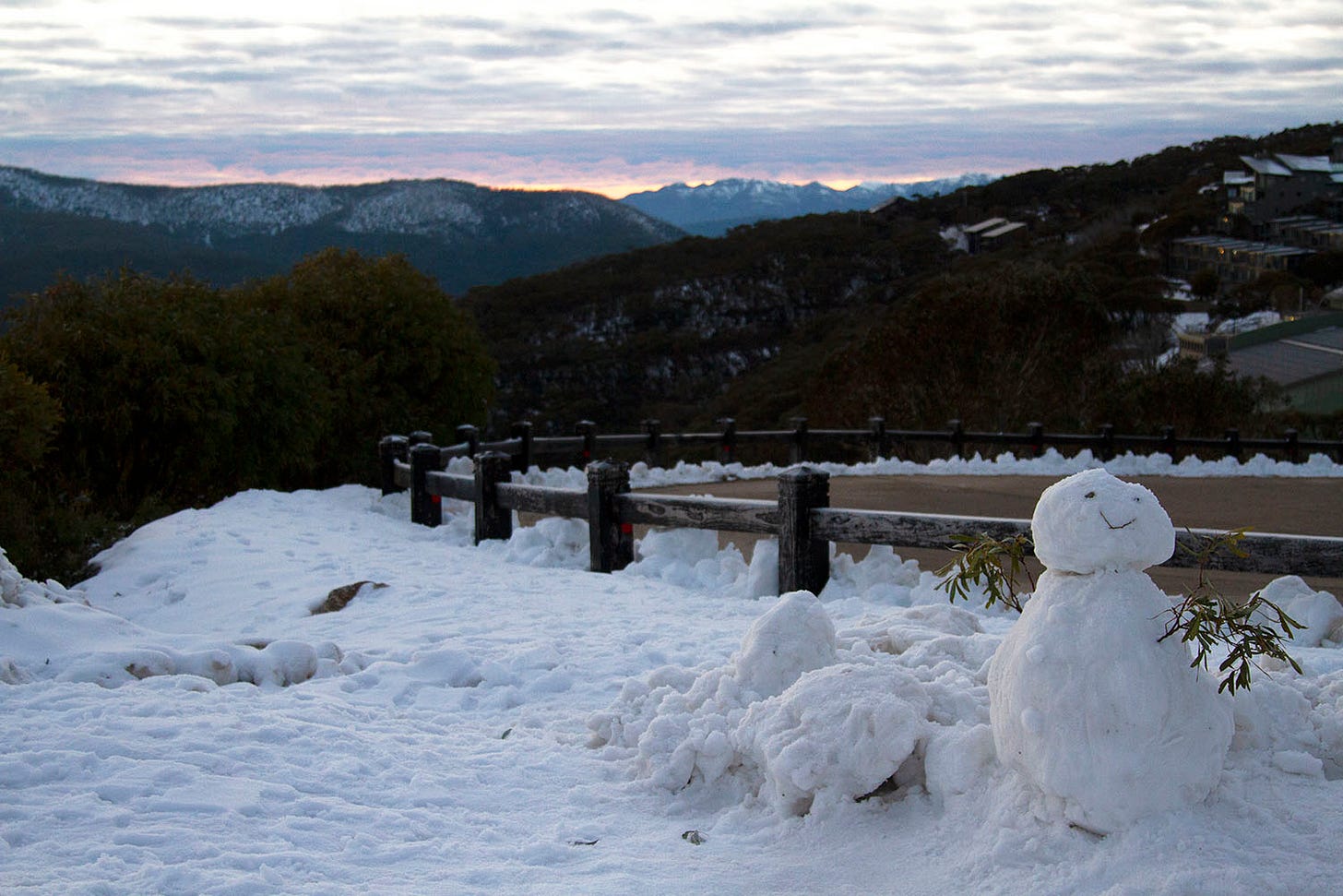 A small snowman by the side of the road on top of a mountain, with snow covered mountains in the distance.