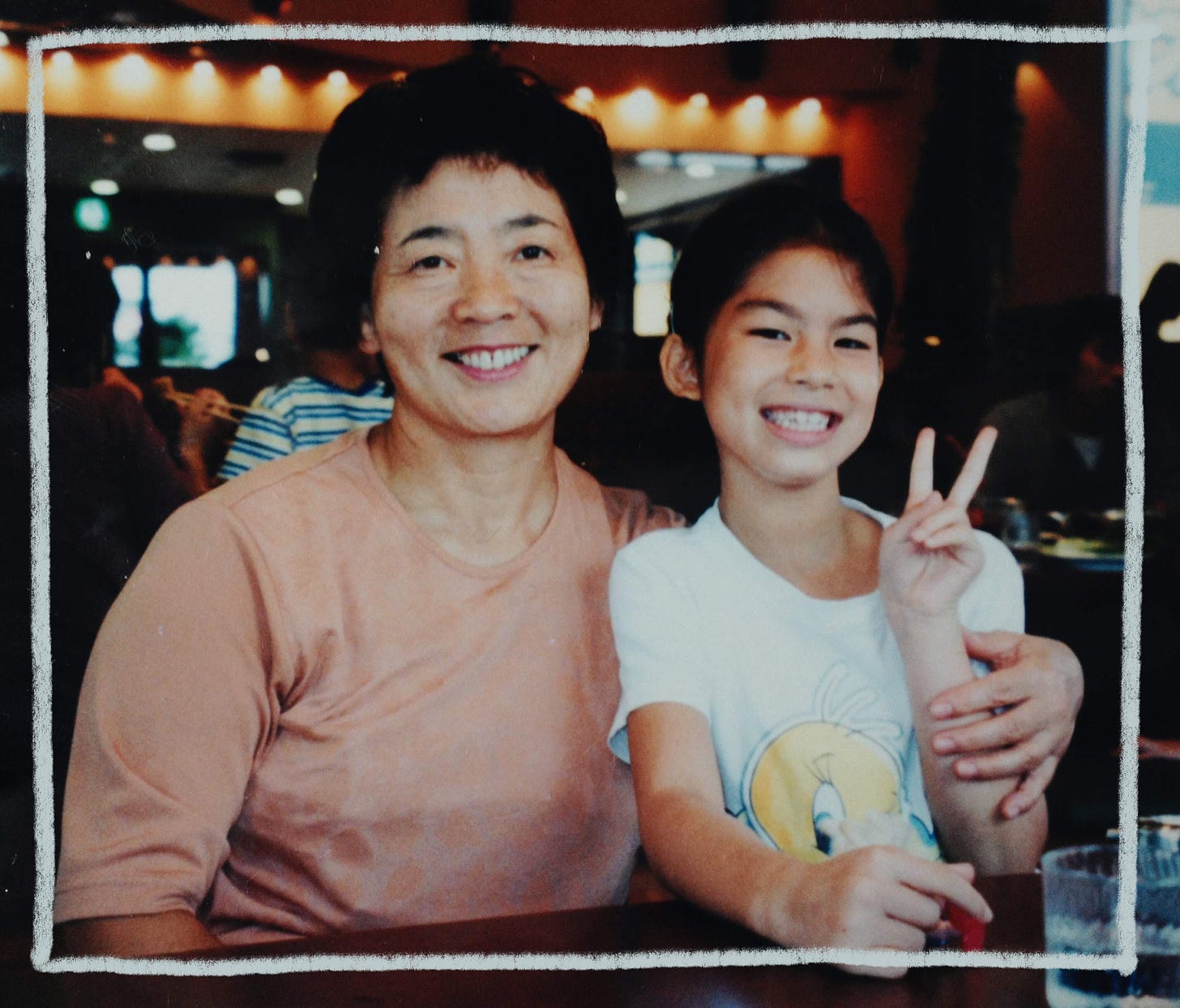 An old photo of the author and Obaachan, her Japanese grandma. They are sitting together at a restaurant, smiling for the camera. Obaachan has her left arm around Erika, and Erika is holding up a peace sign.