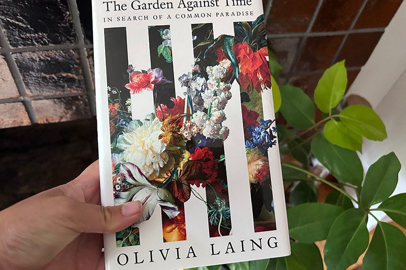 Hand holding a copy of 'The Garden Against Time: In Search of a Common Paradise by Olivia Laing' in front of a houseplant.