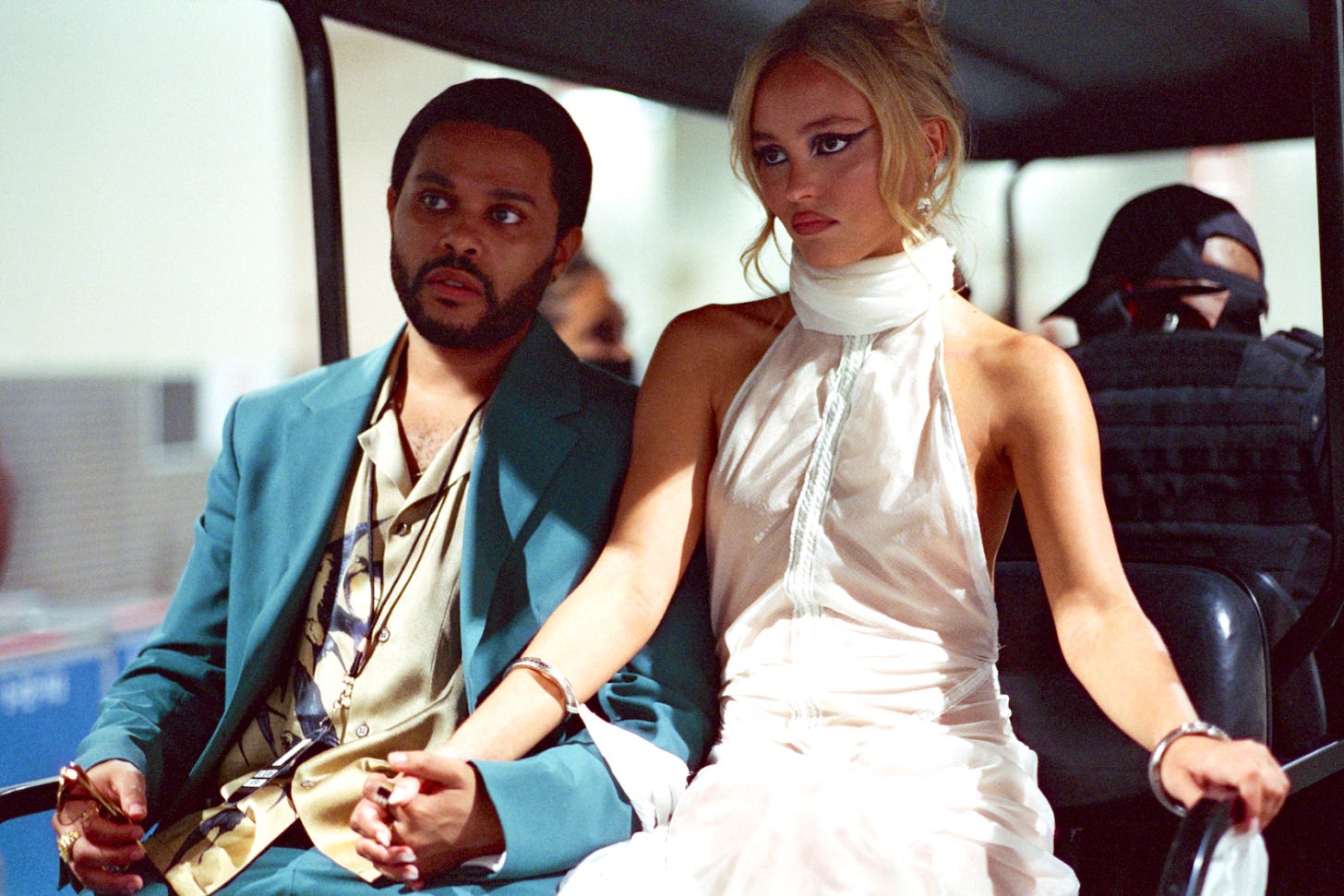 TV still from The Idol. The Weeknd and Lily-Rose Depp's characters hold hands on a bus, dressed in fancy clothes in the sunlight.