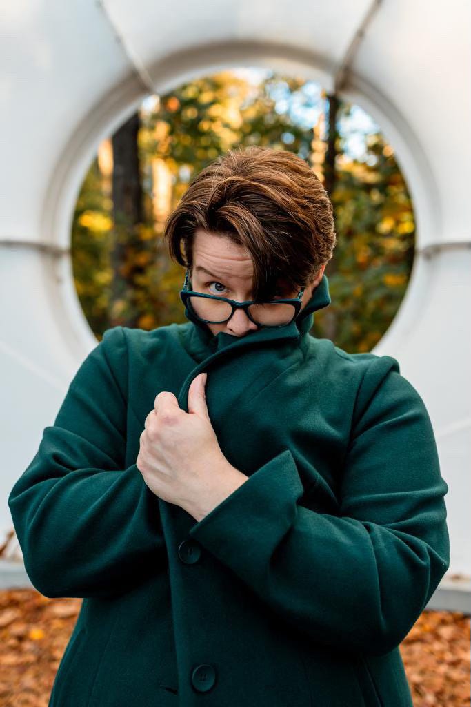 Nonbinary human with white skin and short brownish hair looks with raised eyebrow over green glasses while holding face in green coat. Buckminster Fuller's Fly Eye Dome frames their head from behind.