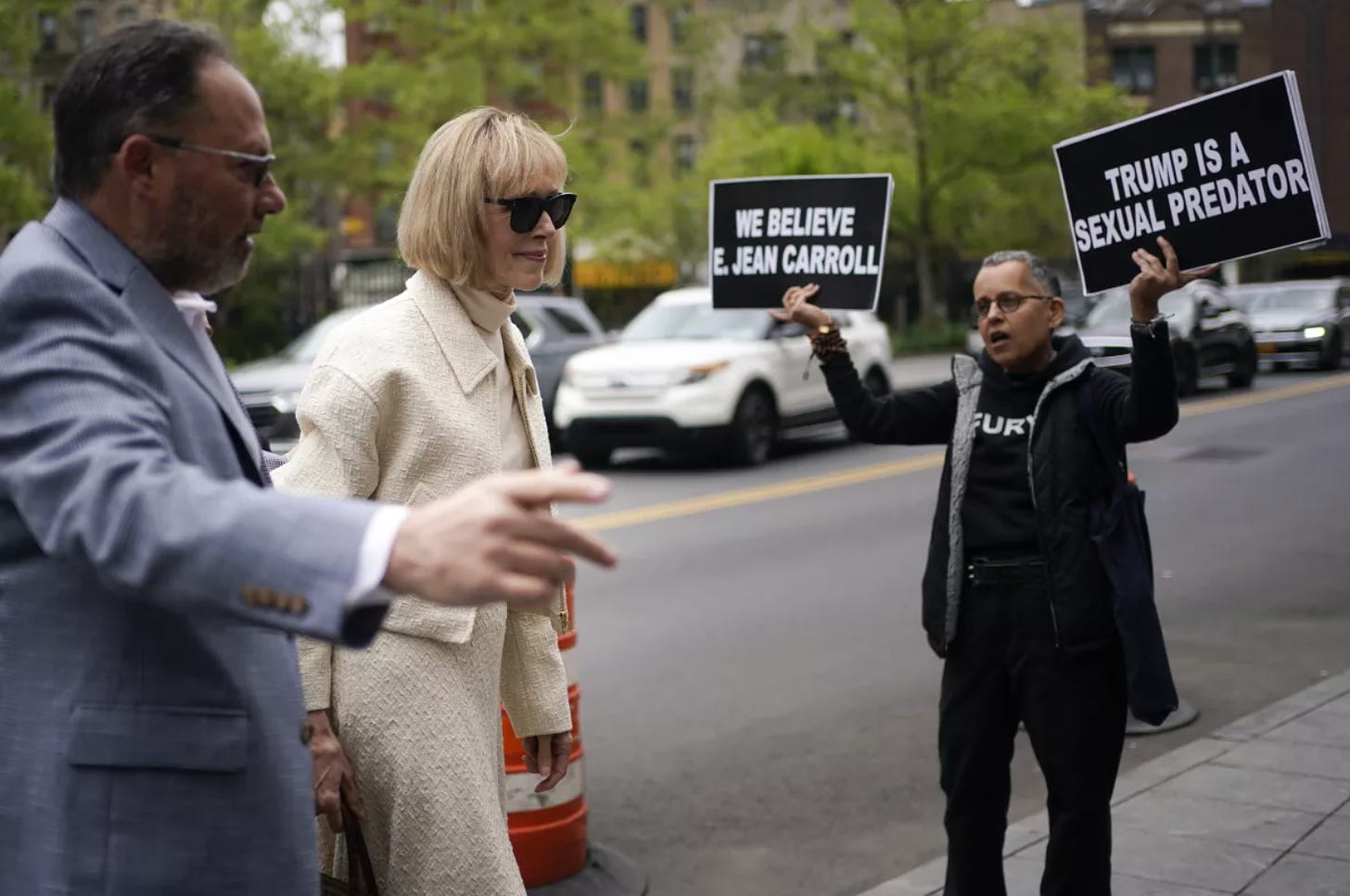 E. Jean Carroll walking into court. Behind her is a person holding up one sign that says "We believe E. Jean Carroll" and another sign that says "Trump is a sexual predator."