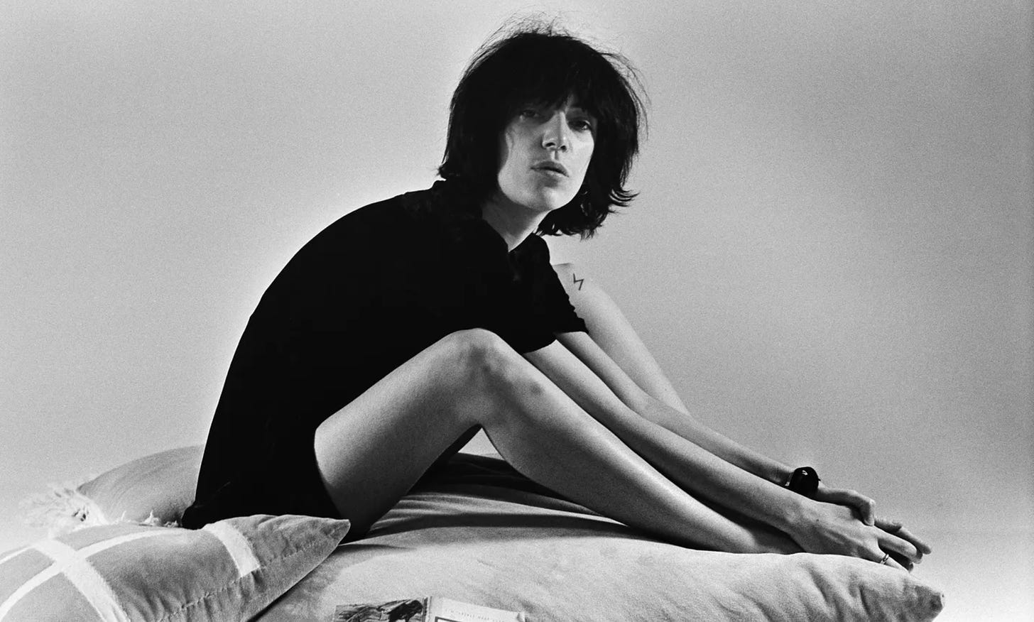 Patti Smith jovem, sentada numa cama de lado, olhando para a câmera. Está vestida apenas com uma camiseta preta, tem cabelos nos ombros e está com o corpo curvado em direção aos pés. Foto p&b