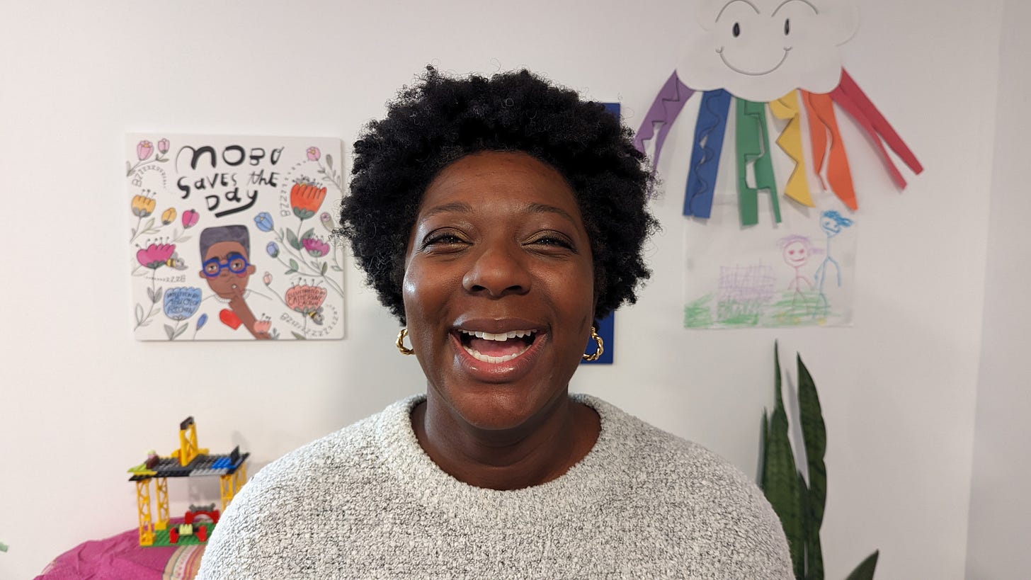 Abiola Regan, a woman smiling energetically at the camera, her background wall plastered with kids photos and the cover of her children's book Mobo Saves the Day