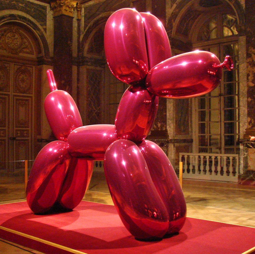 Estátua de uma cachorro feito de balão: ela é rosa, brilhante e reflexiva, parecendo de fato um balão de verdade. Ela está sob um tapete vermelho e um recinto grande e luxuoso, parecendo um prédio antigo, como um museu ou castelo. Ela está em exposição.