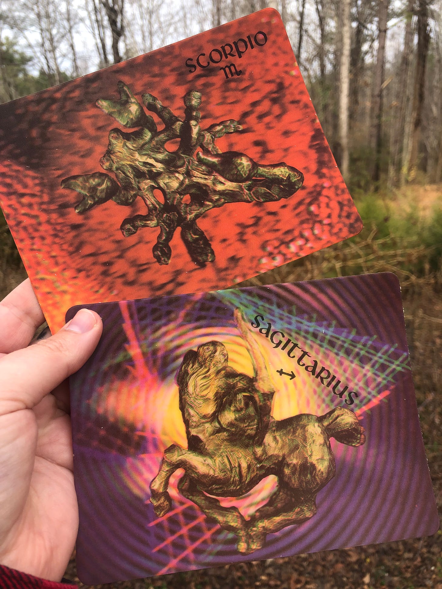 a white hand holds two holographic postcards up against the woods outside in autumn. the top postcard is red with a gold metallic scorpion figure on it with the text “scorpio” and glyph for the zodiac sign of scorpio. the bottom postcard is a background of purple with black radial circles. there are grid like lines spiraling out from the center in pink blue and green. the center image is of a golden figure of the centaur archer which represents the sign sagittarius. text reads “sagittarius” with the glyph for the zodiac sign sagittarius. 