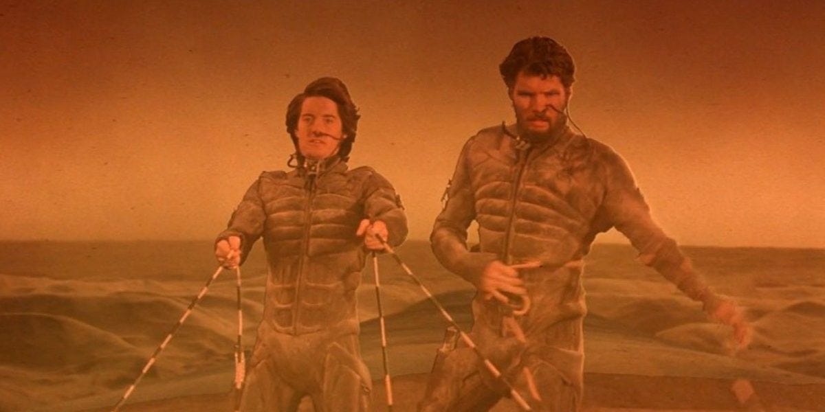 Kyle MacLachlan as Paul Atreides rides a worm at the end of David Lynch's Dune. The scene is very orange. Paul holds the reigns of the worm. Stillgar stands beside him.