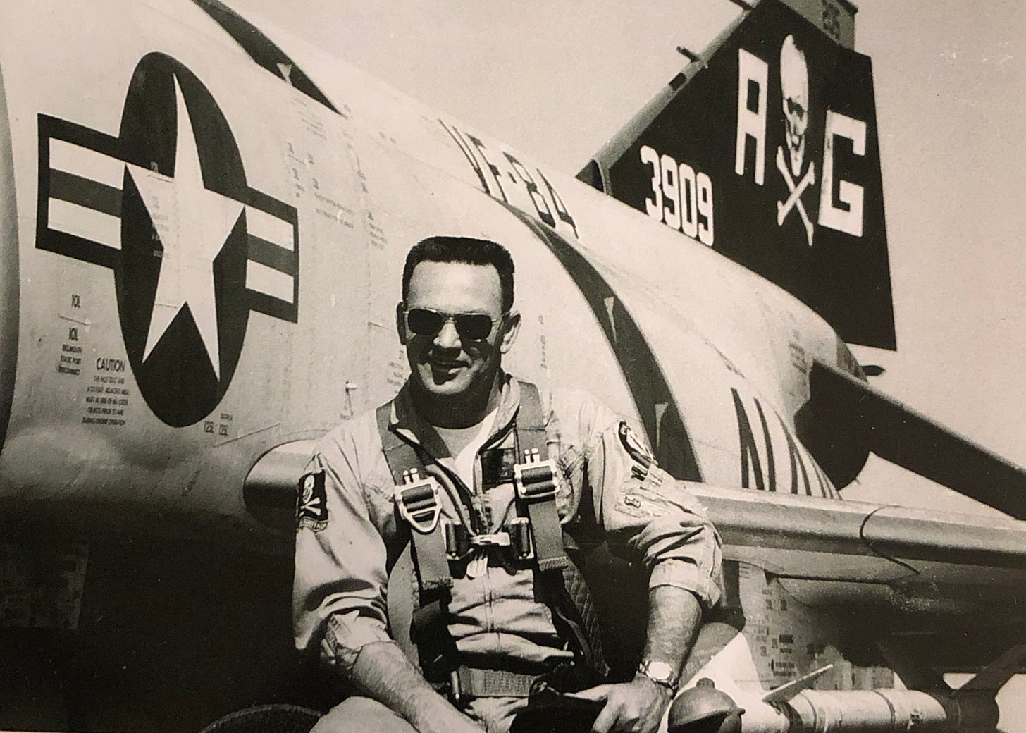 A man with a crewcut wearing a flightsuit and aviator shades poses in front of a Navy jet.