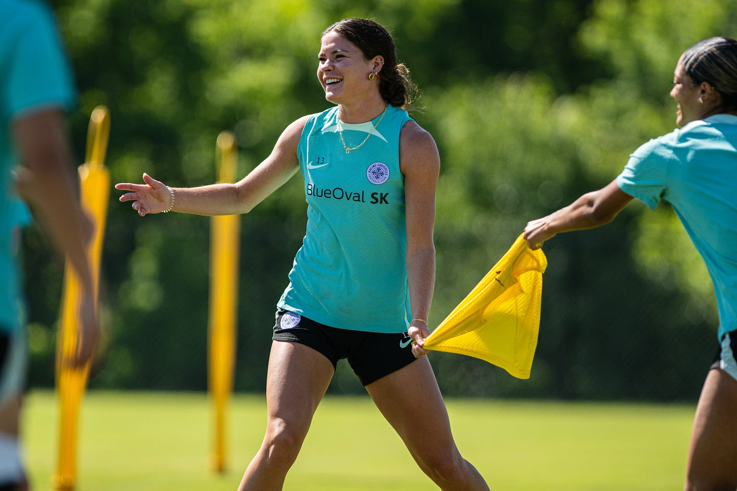 A player in a teal top and black shorts stands in the center facing forward laughing as a teamate pulls on the yellow cloth she's holding