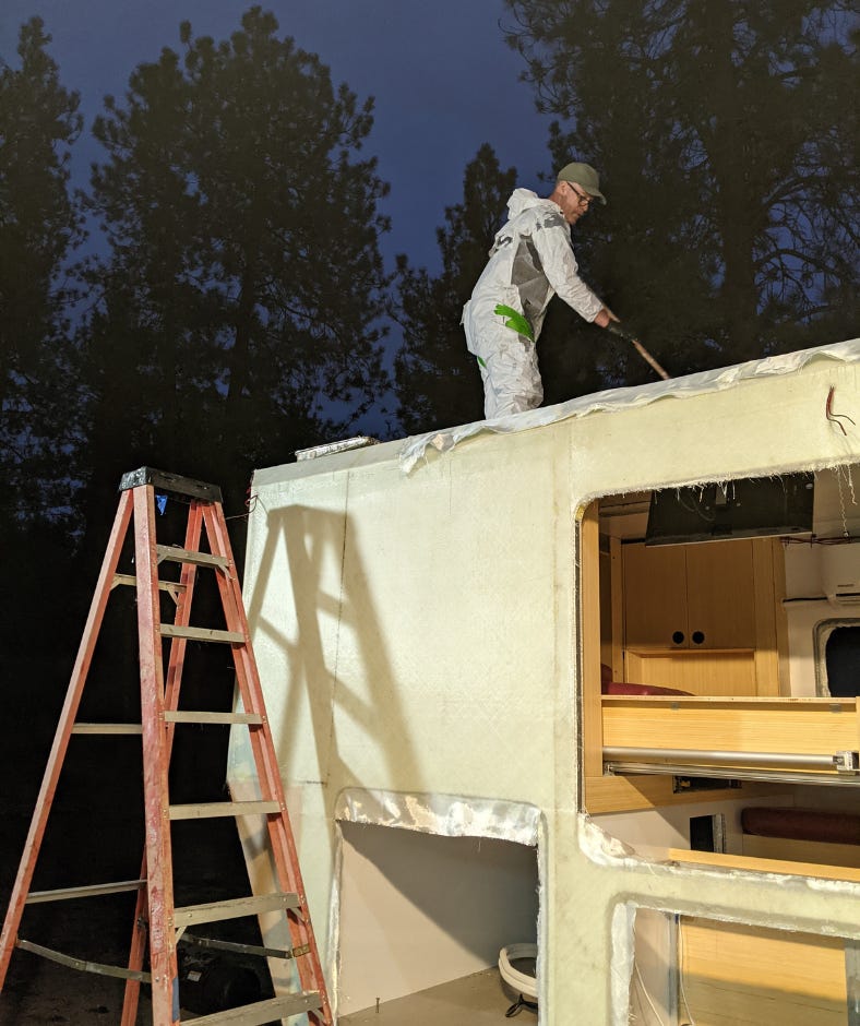 man standing on roof of habitation box, rolling more epoxy over more fiberglass, but this time against a dark night sky with floodlights