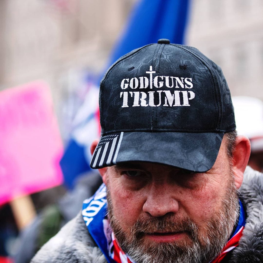 A man wears a hat that reads \"God Guns Trump\" with a cross.
