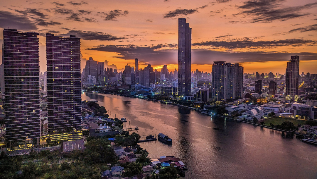 View of Bangkok at sunrise with the Chao Phraya River in the foreground. The sky is purple and orange. 