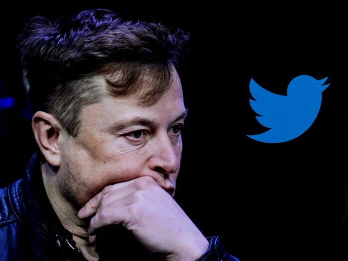 Elon Musk holds his hand to his chin against a black background with the blue Twitter logo