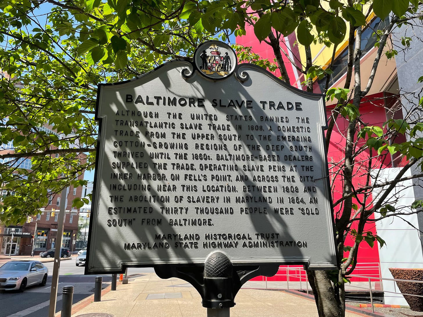 A “BALTIMORE SLAVE TRADE” historic marker standing on a street, with trees and traffic in the background. The marker reads: “ALTHOUGH THE UNITED STATES BANNED THE TRANSATLANTIC SLAVE TRADE IN 1808, A DOMESTIC TRADE FROM THE UPPER SOUTH TO THE EMERGING COTTON-GROWING REGIONS OF THE DEEP SOUTH THRIVED UNTIL THE 1860s. BALTIMORE-BASED DEALERS SUPPLIED THE TRADE, OPERATING SLAVE PENS AT THE INNER HARBOR, ON FELL’S POINT, AND ACROSS THE CITY, INCLUDING NEAR THIS LOCATION. BETWEEN 1808 AND THE ABOLITION OF SLAVERY IN MARYLAND IN 1864, AN ESTIMATED THIRTY THOUSAND PEOPLE WERE ‘SOLD SOUTH’ FROM BALTIMORE.”