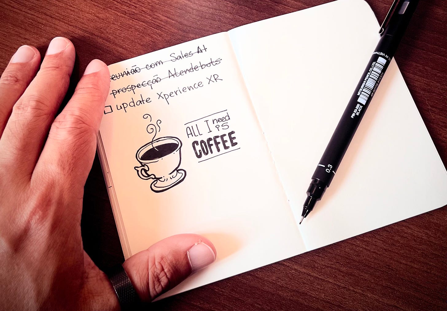  A person's hand rests on a wooden desk next to an open notebook displaying handwritten notes and a whimsical drawing of a coffee cup with the phrase "ALL I need IS COFFEE." A fine-tip black ink pen lies adjacent to the notebook.