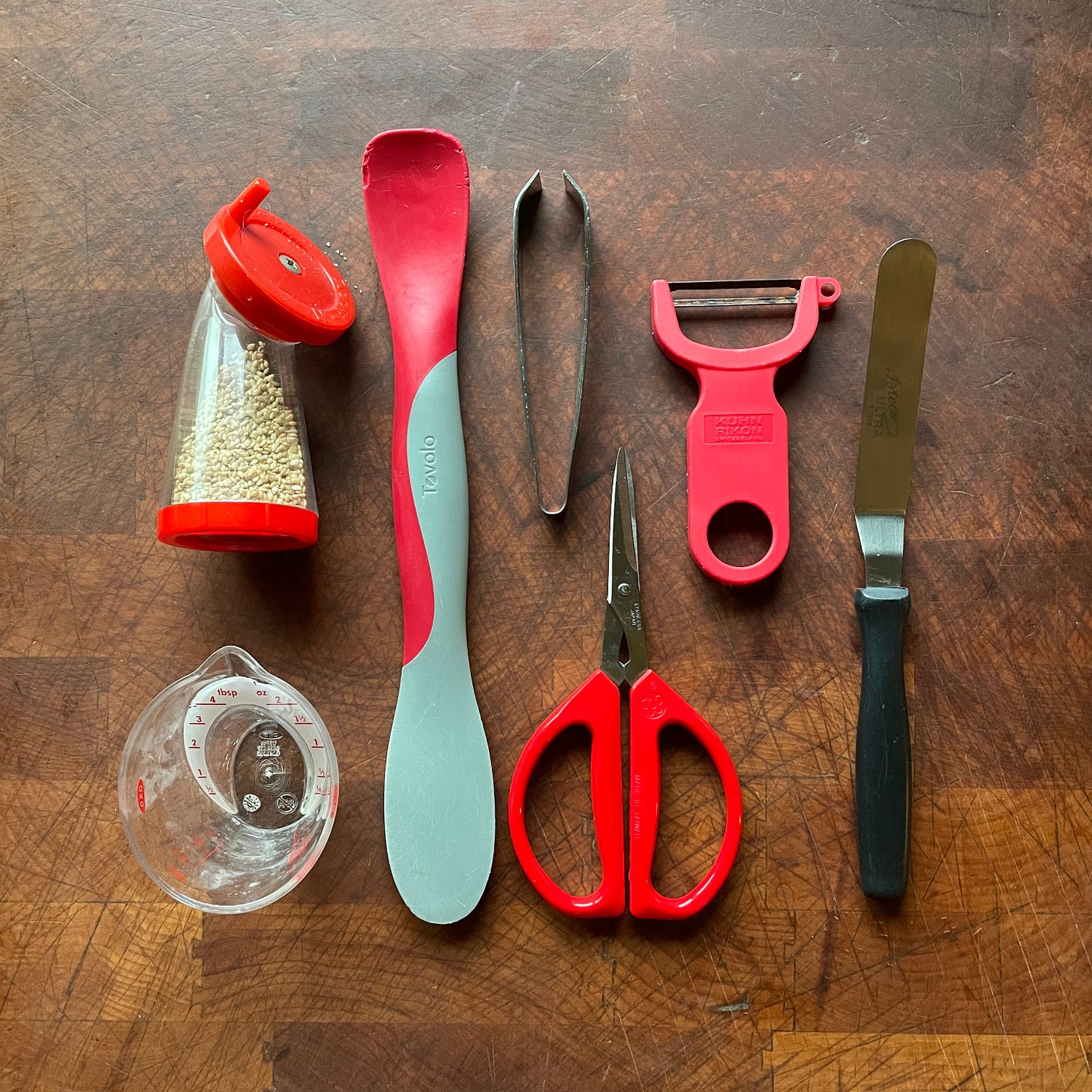 Overhead photo of small cooking tools including a 1/4-cup plastic measuring cup, a jar scraper, a sesame seed grinder, a small pair of red-handled scissors, fish tweezers, a red vegetable peeler, and a small offset spatula with a black plastic handle