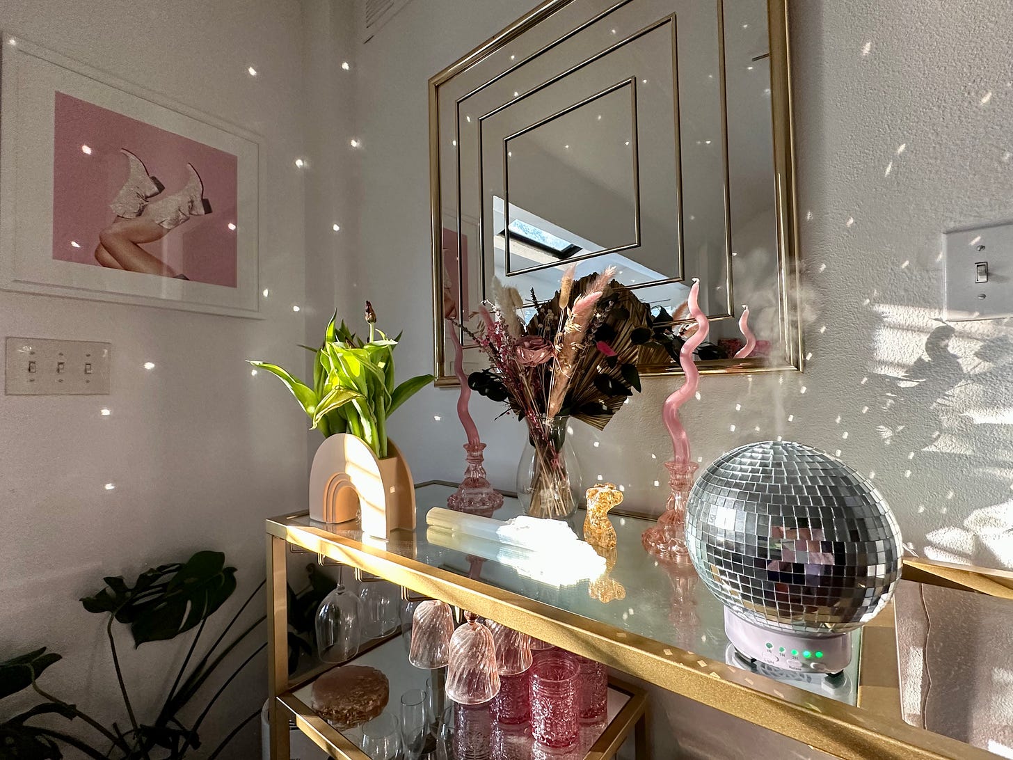 A vase with flowers, a clear crystal, two pink curvy candlesticks, and s disco ball oil diffuser sit on top of a mirrored bar cart with gold edges in a living room with a square golden mirror and an image of a woman kicking up cowboy boots on the wall.