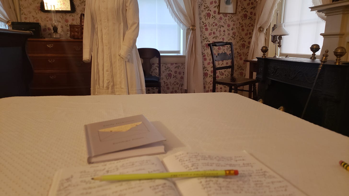 View from the studio session writing table of an open notebook filled with writing in pencil. A yellow-green pencil is laid across the pages. A gray hardcover book of envelope poems is on the table. In the background, the form with Dickinson's white dress, her dresser, one of the windows, and to the right her small table and chair set in front of another window, with the fireplace to the right of the table. 