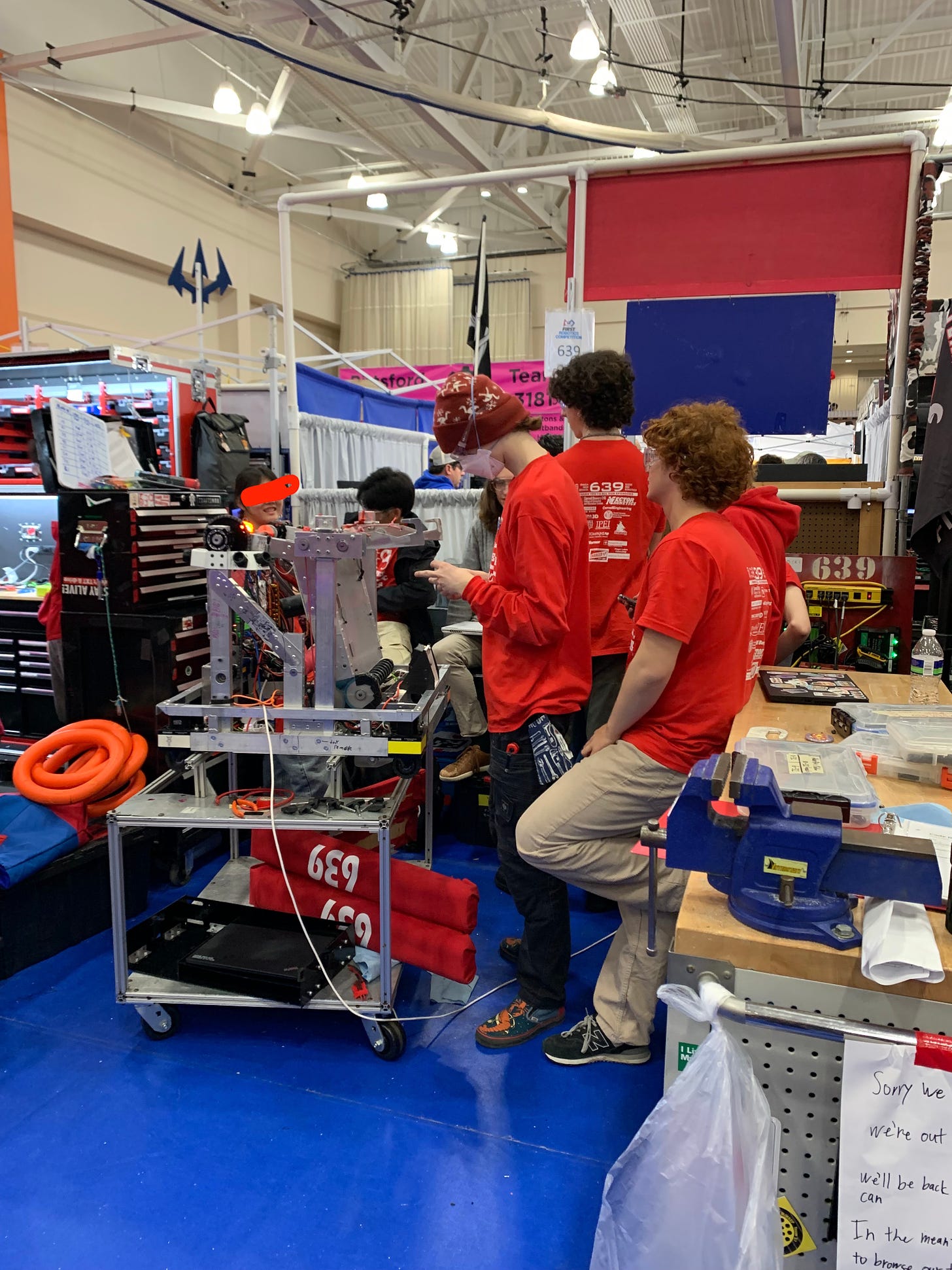 high school students in red t-shirts stand around a robot in the middle of a bustling arrangment of booths full of students fixing robots