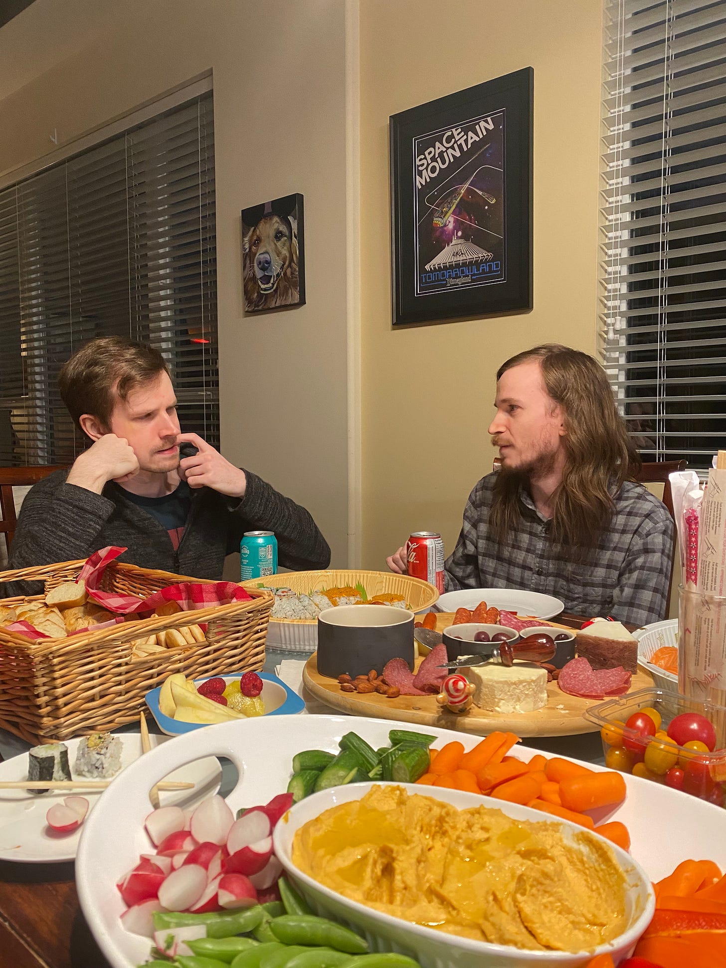 In the foreground, a plate of crudités and sweet potato hummus, surrounded by picked-at platters of charcuterie and sushi, and a basket of bread and crackers. On the far end of the table, Ken (long-hair, a short beard, and a grey checkered shirt) sits on the right talking to Trav on the left (short hair and wearing a grey sweater), who has a look of concentration on his face, his face resting on his hand.