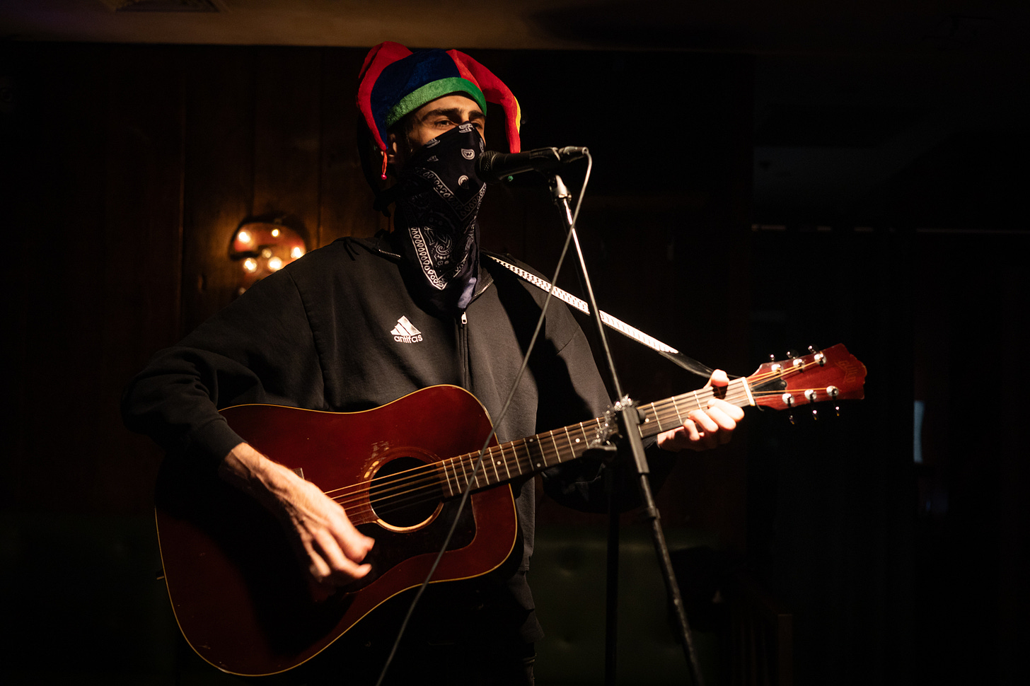 jester of no court performs with jester hat, acoustic guitar, and black bandana face mask at queers n peers variety show